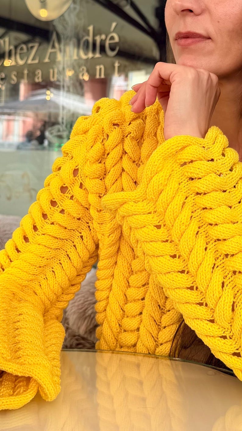 May include: A bright yellow long-sleeve sweater featuring a prominent chunky cable knit pattern with vertical braided textures. The garment has a thick, textured weave throughout the sleeves and body, showcasing a vibrant, solid yellow color.