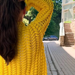 May include: A bright yellow long-sleeve cardigan featuring a prominent vertical cable knit pattern with openwork detailing between the stitches. The garment has a relaxed fit with long sleeves and a textured surface created by the thick yarn construction.