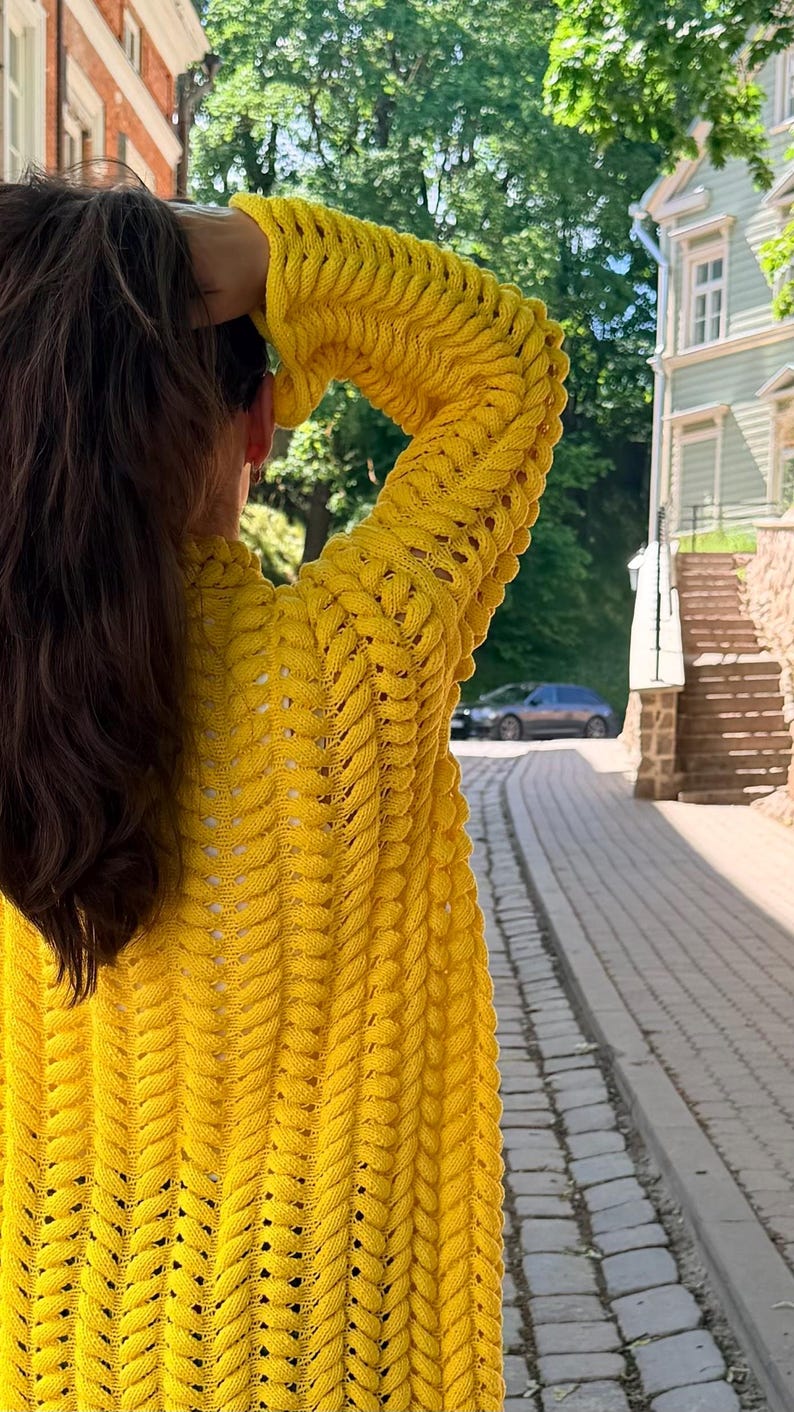 May include: A bright yellow long-sleeve cardigan featuring a prominent vertical cable knit pattern with openwork detailing between the stitches. The garment has a relaxed fit with long sleeves and a textured surface created by the thick yarn construction.