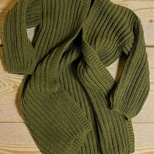 May include: Olive green knit cardigan with a ribbed texture. The long-sleeved sweater is draped over a wooden surface, showcasing its open front and cozy design. The garment is made of a thick, warm material.