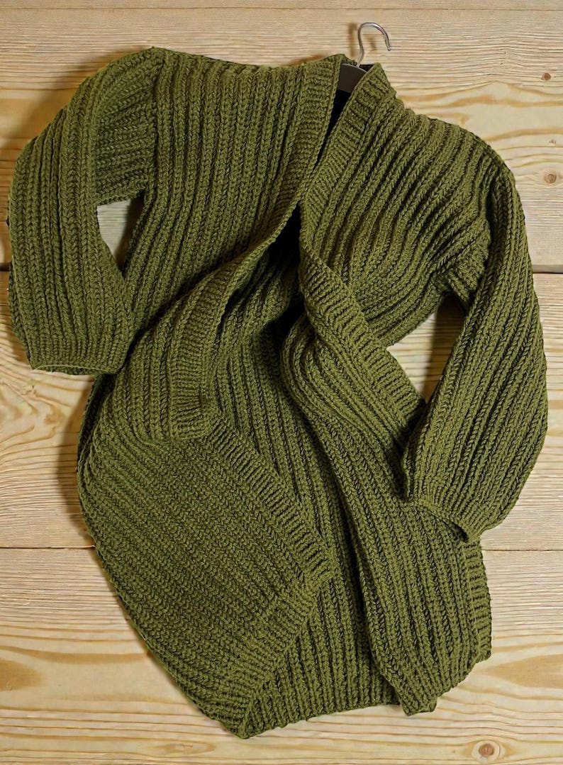 May include: Olive green knit cardigan with a ribbed texture. The long-sleeved sweater is draped over a wooden surface, showcasing its open front and cozy design. The garment is made of a thick, warm material.