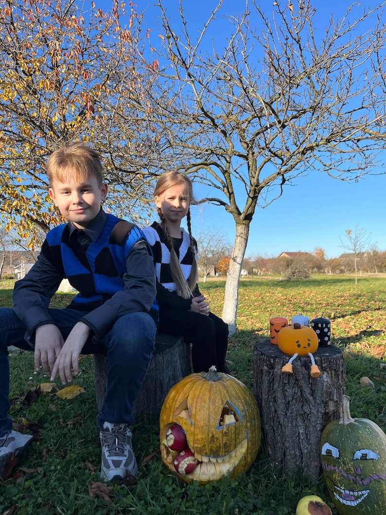 May include: Two children sit on a tree stump in a sunny outdoor scene. A carved pumpkin with a smiling face and apples inside sits in front of them. Other carved pumpkins and a pumpkin-shaped plush toy are nearby, suggesting a Halloween theme.