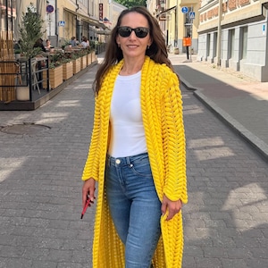 May include: A bright yellow, long-length, chunky knit cardigan featuring a prominent braided stitch pattern, worn over a plain white t-shirt and blue denim jeans, paired with white sneakers and dark-tinted sunglasses for a casual look.