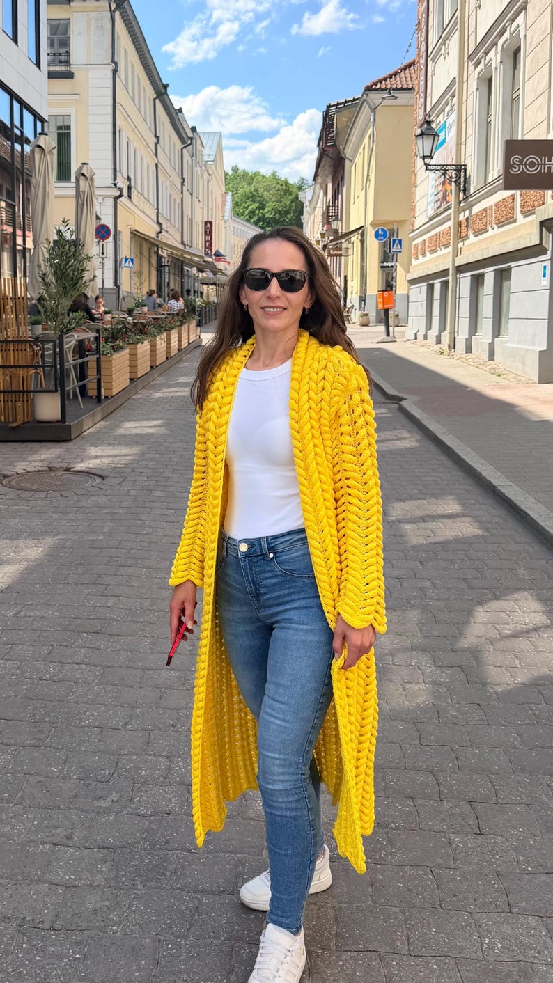 May include: A bright yellow, long-length, chunky knit cardigan featuring a prominent braided stitch pattern, worn over a plain white t-shirt and blue denim jeans, paired with white sneakers and dark-tinted sunglasses for a casual look.