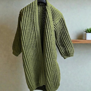 May include: Olive green, open-front cardigan with a ribbed knit texture. The longline sweater features three-quarter sleeves and hangs from a black hanger. The garment is displayed against a neutral background, showcasing its simple, yet stylish design.