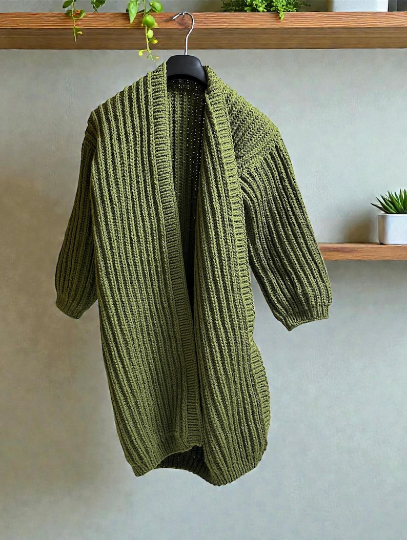 May include: Olive green, open-front cardigan with a ribbed knit texture. The longline sweater features three-quarter sleeves and hangs from a black hanger. The garment is displayed against a neutral background, showcasing its simple, yet stylish design.
