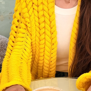 May include: A bright yellow open-front cardigan featuring a chunky cable knit texture with vertical braided patterns and long sleeves. The garment is worn over a plain white top. A white ceramic cup with a red logo and latte art is visible.