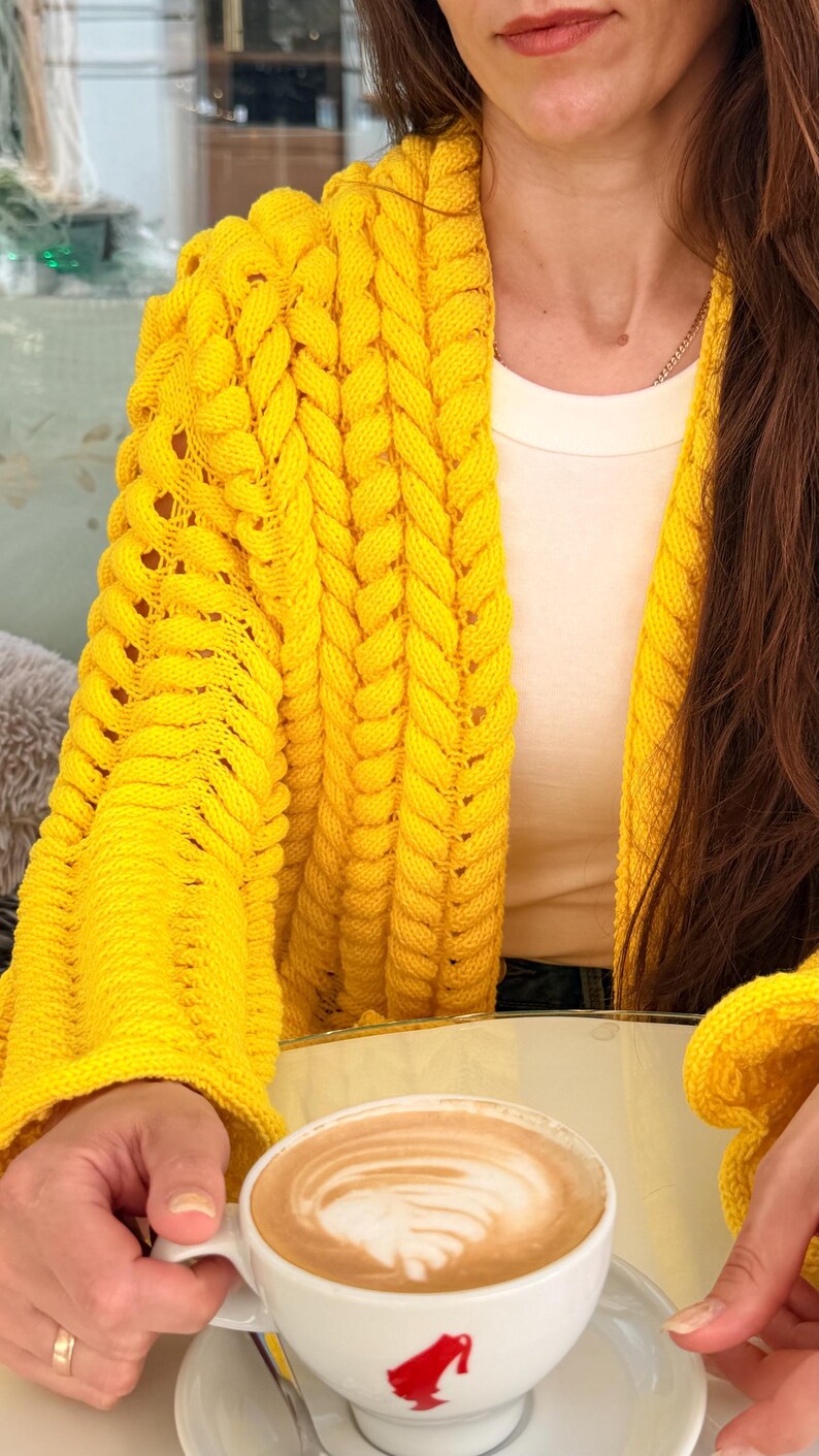 May include: A bright yellow open-front cardigan featuring a chunky cable knit texture with vertical braided patterns and long sleeves. The garment is worn over a plain white top. A white ceramic cup with a red logo and latte art is visible.