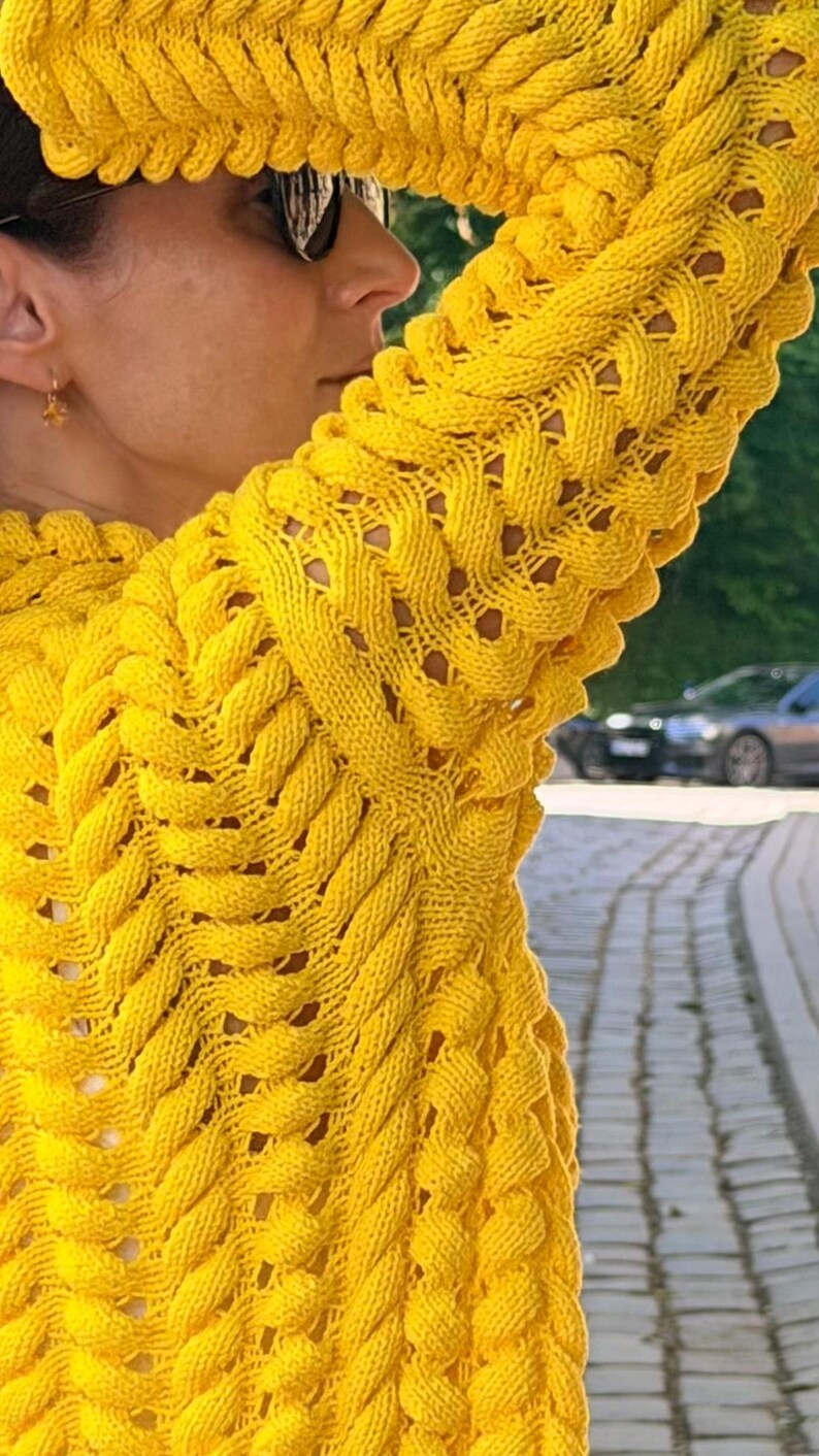 May include: A bright yellow long-sleeve sweater featuring a prominent, raised cable knit pattern that runs vertically along the sleeves and body. The knit construction creates a textured, braided appearance with small open spaces between the thick yarn.