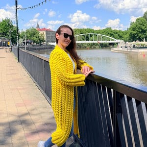 May include: A long, bright yellow cardigan featuring a chunky, braided knit pattern with long sleeves and an open front design. The garment is worn over a white top and blue denim jeans, paired with white sneakers and dark sunglasses.