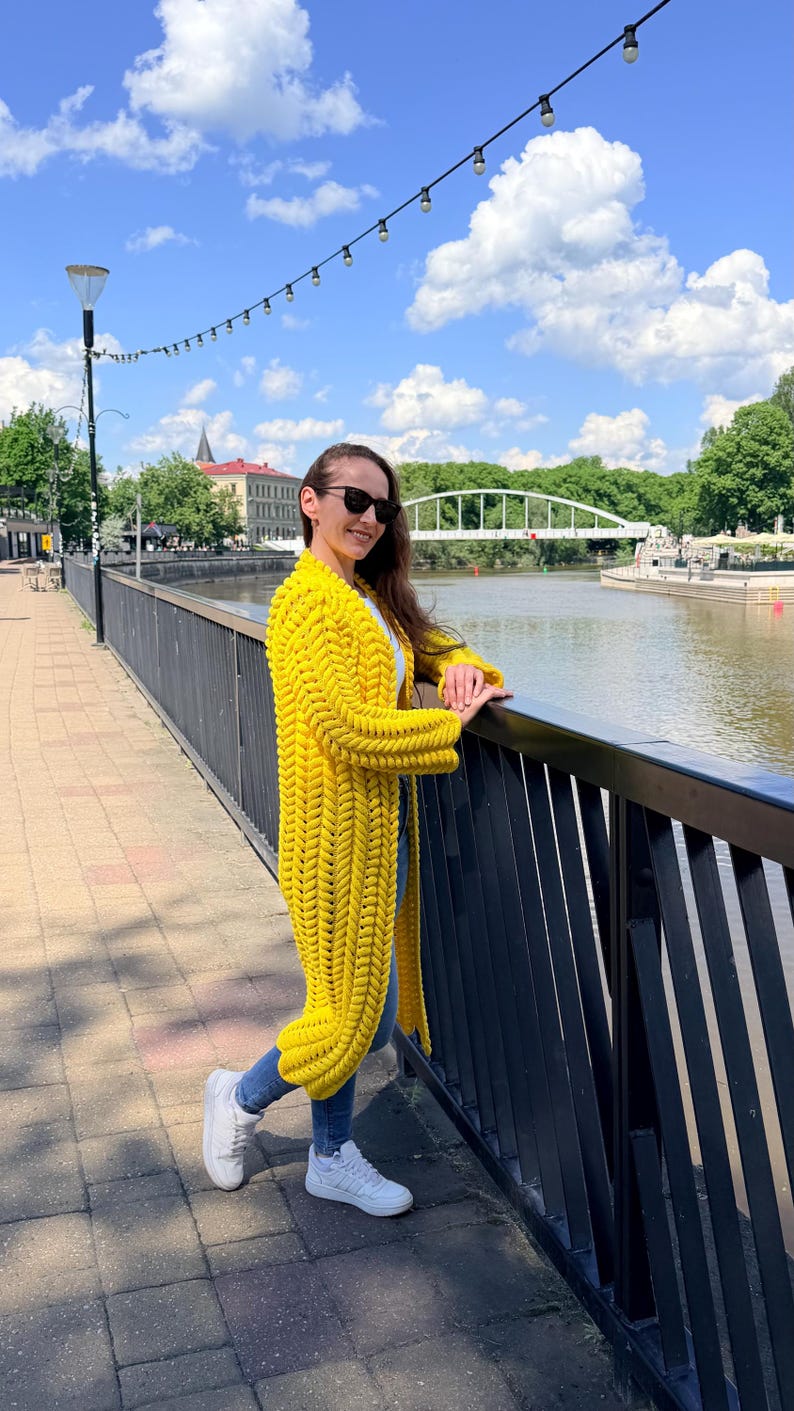 May include: A long, bright yellow cardigan featuring a chunky, braided knit pattern with long sleeves and an open front design. The garment is worn over a white top and blue denim jeans, paired with white sneakers and dark sunglasses.