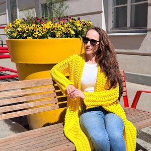 May include: A bright yellow chunky knit cardigan with a prominent braided texture, worn over a plain white t-shirt and blue denim jeans, paired with white sneakers and black sunglasses, creating a vibrant and casual layered outfit ensemble.