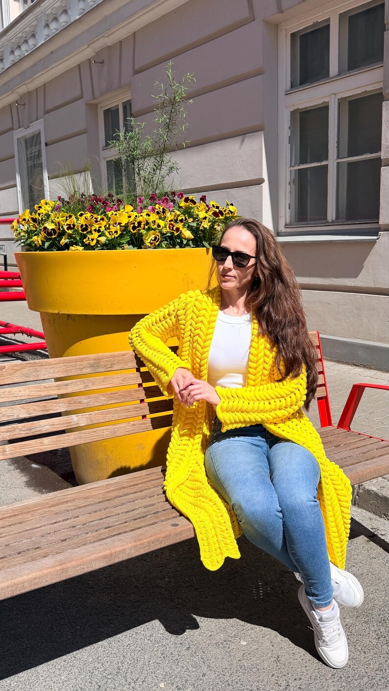 May include: A bright yellow chunky knit cardigan with a prominent braided texture, worn over a plain white t-shirt and blue denim jeans, paired with white sneakers and black sunglasses, creating a vibrant and casual layered outfit ensemble.
