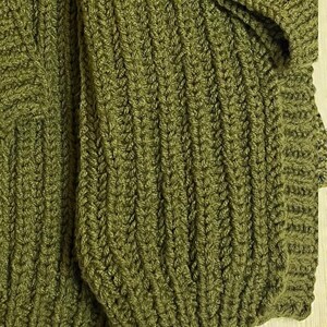 May include: Close-up of a knitted garment in a muted olive green color. The fabric has a textured, ribbed pattern, with visible stitches creating a detailed design. The image shows a portion of the garment, possibly a sweater or cardigan.