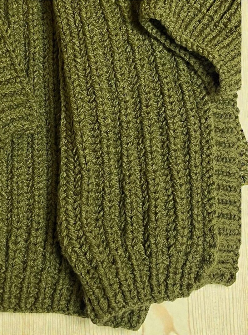 May include: Close-up of a knitted garment in a muted olive green color. The fabric has a textured, ribbed pattern, with visible stitches creating a detailed design. The image shows a portion of the garment, possibly a sweater or cardigan.