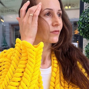 May include: A bright yellow knit cardigan featuring a prominent braided cable stitch pattern along the front panels and sleeves. The garment is worn over a simple white shirt, showcasing a thick, textured yarn construction with long sleeves.