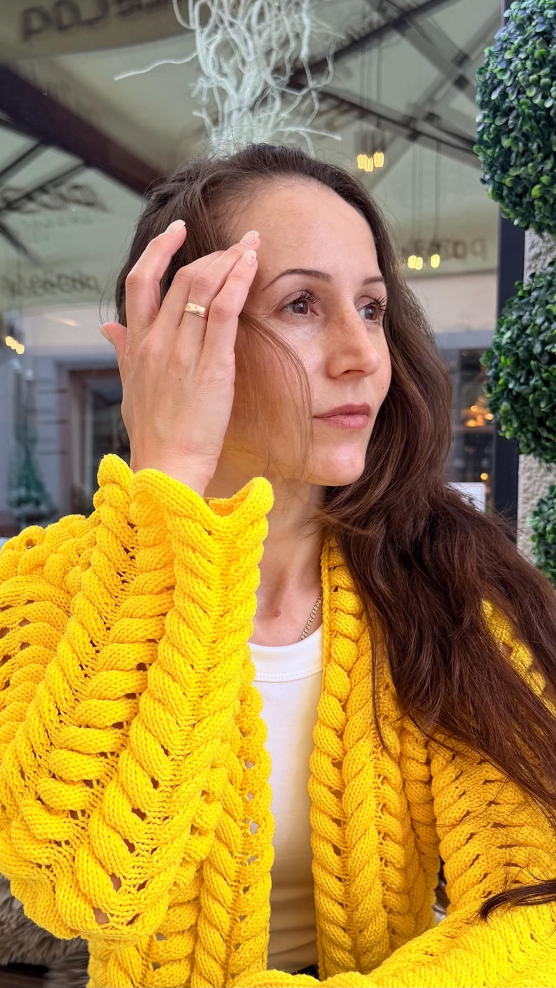 May include: A bright yellow knit cardigan featuring a prominent braided cable stitch pattern along the front panels and sleeves. The garment is worn over a simple white shirt, showcasing a thick, textured yarn construction with long sleeves.