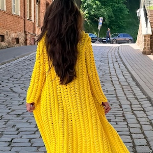 May include: A bright yellow, long-sleeved, open-front cardigan featuring a chunky, vertical braided knit pattern. The garment reaches below the knees and is worn over blue denim jeans and white sneakers, showcasing a textured, heavy yarn construction.