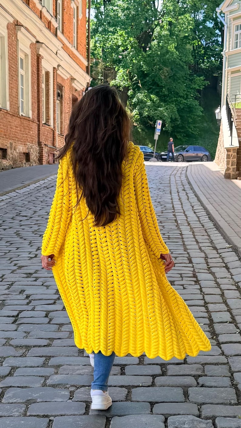 May include: A bright yellow, long-sleeved, open-front cardigan featuring a chunky, vertical braided knit pattern. The garment reaches below the knees and is worn over blue denim jeans and white sneakers, showcasing a textured, heavy yarn construction.