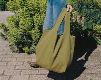 May include: Olive green tote bag with long handles. The bag is made of a soft, durable fabric and is being held by a person wearing blue jeans and a green sweater. The bag is large and appears to be empty. The background includes green foliage and a brick walkway.