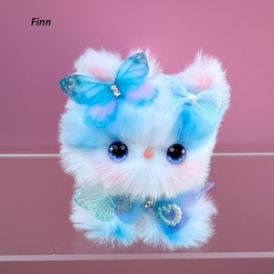 May include: A soft, fluffy cat-like toy in blue, white, and pink hues. It has large, detailed eyes, and is adorned with a butterfly and bow. The name "Finn" is written in the top left corner.