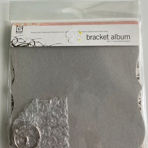 Peut inclure: Un album en carton gris avec un design en forme de crochet et une reliure à anneau unique. L'album est emballé dans un sac en plastique transparent avec un fond à motifs blanc et noir.