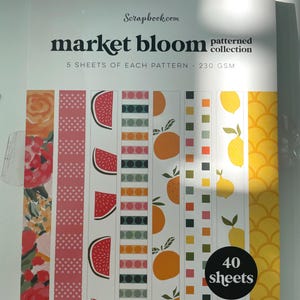 May include: A scrapbook paper pad titled "market bloom patterned collection" with various fruit and floral patterns. The patterns include watermelon slices, oranges, lemons, and floral designs. The pad contains 40 sheets.
