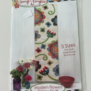 Op de afbeelding: Een naai-patroon voor een moderne smalle bloemenquilt. Het patroon bevat instructies voor het maken van een wandquilt, een tafelkleed of een bedsprei. De quilt heeft een kleurrijk bloemenontwerp op een witte achtergrond.