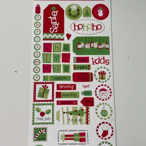 Op de afbeelding: Vel kerst-thema kartonnen stickers. Ontwerpen omvatten de kerstman, kerstbomen, cadeaudozen en de woorden "Christmas", "Holly Jolly" en "dec. 25". Rode, groene en witte kleuren.