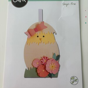 May include: A Sizzix Thinlits die set package, model 664362 by Georgie Evans, featuring 12 metal dies. The packaging displays a paper craft of a yellow chick hatching from a light tan egg, decorated with a pink bow and pink flowers on green grass.