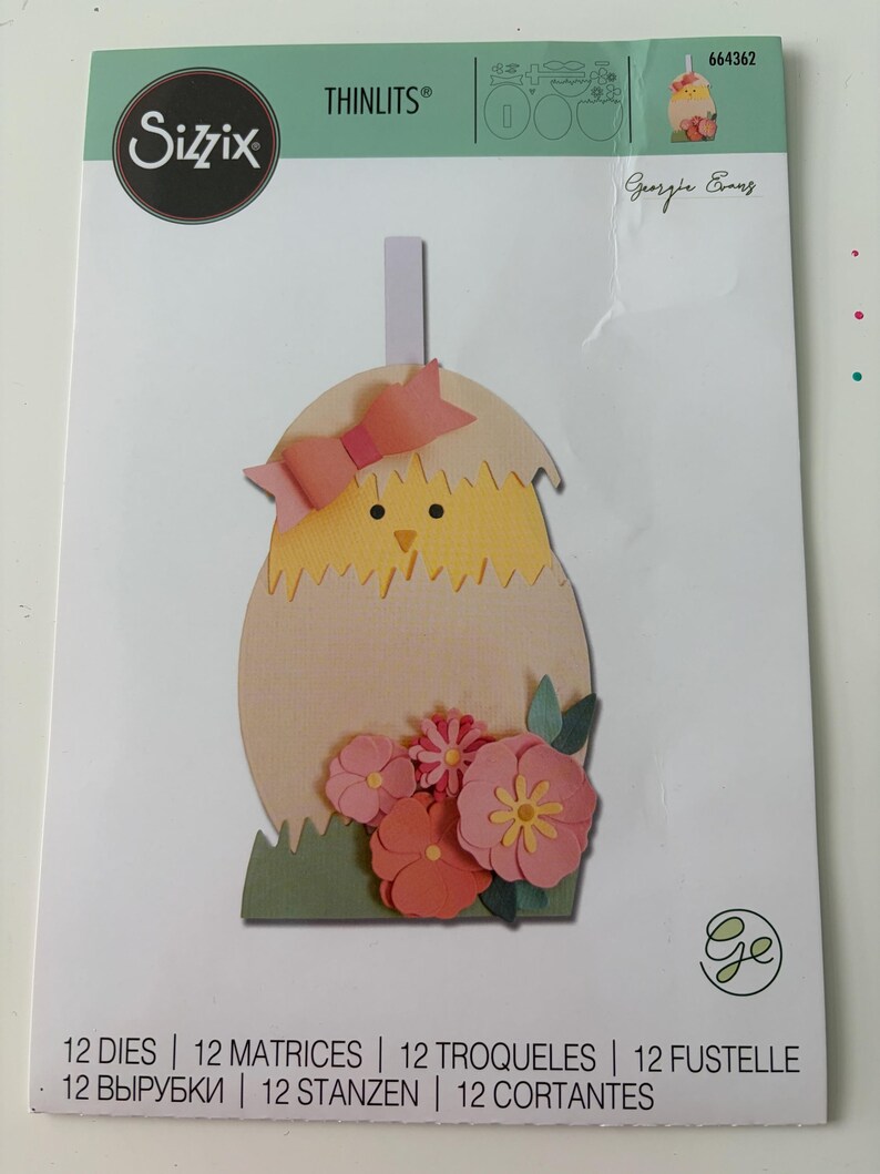 May include: A Sizzix Thinlits die set package, model 664362 by Georgie Evans, featuring 12 metal dies. The packaging displays a paper craft of a yellow chick hatching from a light tan egg, decorated with a pink bow and pink flowers on green grass.