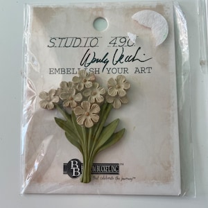 May include: A package containing a floral embellishment. The embellishment features a cluster of small, cream-colored flowers with green leaves and stems. The packaging includes the text "STUDIO 490" and "EMBELISH YOUR ART".