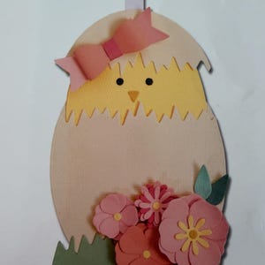 May include: A decorative hanging ornament featuring a yellow chick emerging from a light beige eggshell. The chick wears a pink paper bow on its head. The base of the egg is decorated with layered pink and coral paper flowers and green leaves.