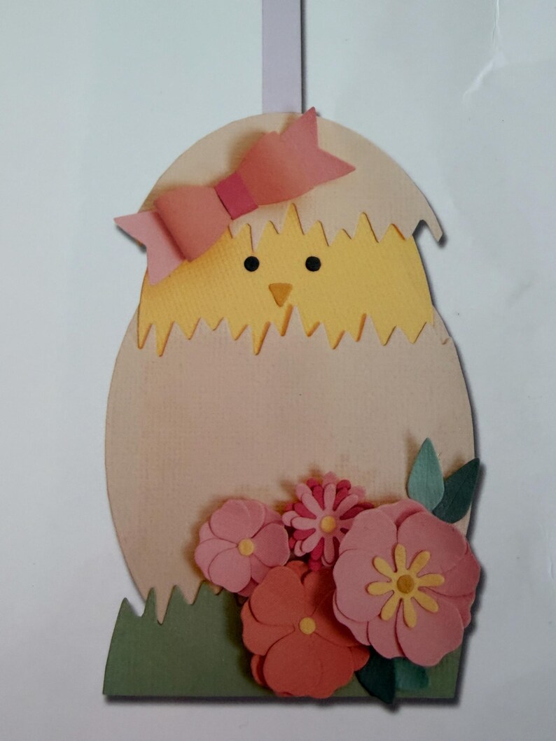 May include: A decorative hanging ornament featuring a yellow chick emerging from a light beige eggshell. The chick wears a pink paper bow on its head. The base of the egg is decorated with layered pink and coral paper flowers and green leaves.