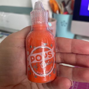May include: A small orange bottle of paint with a white label that says "POPS of Color" and "Scrapbook.com".