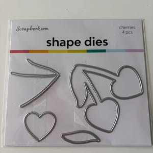 May include: A package of shape dies from Scrapbook.com. The package contains four pieces of cherry-themed dies, including two cherries, a heart, and a leaf. The package has a rainbow stripe design at the bottom.