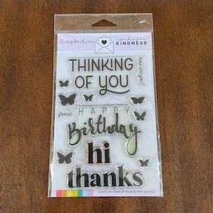May include: A clear plastic package containing a set of clear photopolymer stamps with black ink. The stamps feature various phrases and images, including "Thinking of You", "Happy Birthday", "Hi", "Thanks", and butterflies. The package also includes the text "cards for kindness" and "Scrapbook.com Card Maker Essentials".