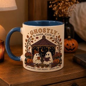 May include: A white ceramic mug with a blue interior and handle, featuring a graphic of two ghosts in a gazebo under a night sky. The text reads GHOSTLY BOOK CLUB at the top and GOOD BOOKS COZY NIGHTS SPOOKY VIBES at the bottom, surrounded by autumn leaves and pumpkins.
