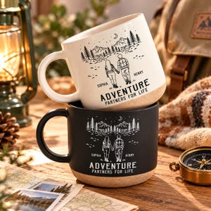 Peut inclure: Deux tasses en céramique, une blanche et une noire, arborant chacune un dessin de deux personnes se tenant la main dans un paysage de montagne avec le texte "SOPHIA", "HENRY" et "ADVENTURE PARTNERS FOR LIFE". Base couleur sable.