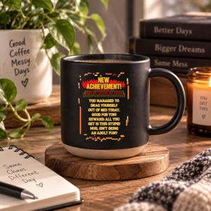 Può includere: Una tazza in ceramica nera con base beige che riporta il testo NEW ACHIEVEMENT! HERE WE FUCKING GO AGAIN! YOU MANAGED TO DRAG YOURSELF OUT OF BED TODAY. GOOD FOR YOU! REWARD: ALL YOU GET IS THIS STUPID MUG. ISN'T BEING AN ADULT FUN?
