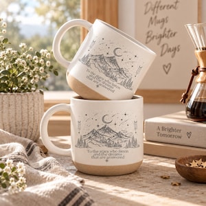 Puede incluir: Dos tazas de cerámica de color crema apiladas verticalmente, con una ilustración de líneas negras de montañas, una luna creciente, estrellas y el texto: To the stars who listen and the dreams that are answered. Cada taza tiene un asa.