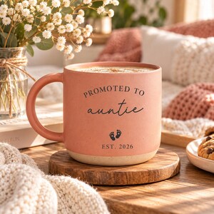 May include: A matte coral-coloured ceramic mug with a rounded handle and a speckled cream-coloured base. The front features black text that reads PROMOTED TO auntie, with a small graphic of two baby footprints and the text EST. 2026 below it.