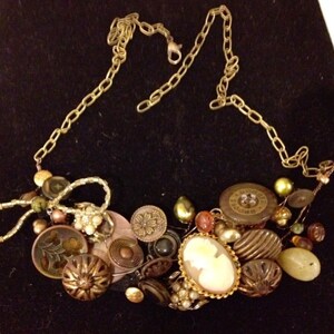 May include: A gold chain necklace with a large cluster of vintage buttons, beads, and other embellishments. The necklace features a variety of colors, including brown, gold, green, and white.