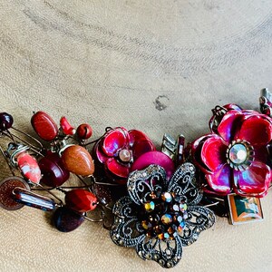 May include: A statement necklace with a mix of red, brown, and black beads, flowers, and metal accents. The necklace features a large, ornate, black metal flower with rhinestones in the center.