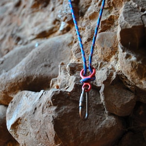 Peut inclure: Une corde rouge et bleue est attachée à un mousqueton argenté avec un clip en plastique rouge. La corde est suspendue à une paroi rocheuse.