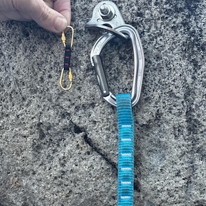 Könnte beinhalten: Ein silbernes Kletter-Absicherungssystem mit einem blau-weißen Gurtband, das daran befestigt ist. Ein goldener Karabiner mit einem schwarz-roten Gurtband ist ebenfalls abgebildet.