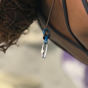 Puede incluir: Primer plano de un mosquetón de escalada y un conector azul sujetos a una cuerda gris. El mosquetón es plateado con un mecanismo de bloqueo dorado. El fondo está borroso.