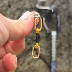 May include: A gold-colored climbing carabiner with a black and yellow cord. The carabiner is held in front of a gray rock wall, with other climbing gear visible in the background.