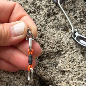 May include: A silver-colored metal clasp with an orange cord and a black rubber sleeve. The clasp is connected to a larger silver metal piece. The image is set against a textured, gray background.