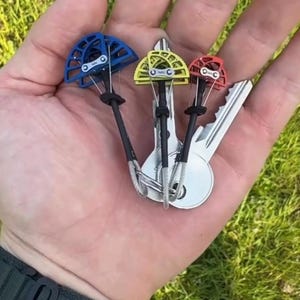 May include: A hand holding three climbing cams in blue, yellow, and red, attached to keys. The cams have a black stem and silver wire. The background is green grass.